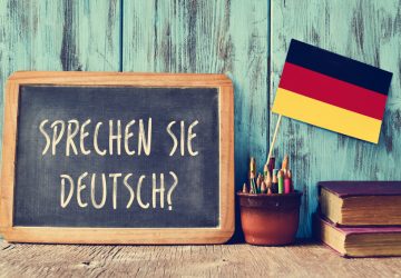 a chalkboard with the question sprechen sie deutsch? do you speak german? written in german, a pot with pencils, some books and the flag of Germany, on a wooden desk
